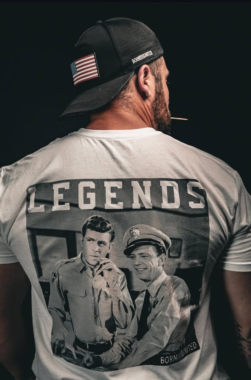 A man in a cap with an American flag and a toothpick faces away from the camera. He wears Born United Private Labels Legend line t-shirt, featuring LEGENDS over a black-and-white image of two men in uniforms—a tribute to timeless American culture. Product: Legends - Andy Griffith.