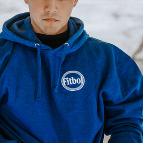 Someone wears the Pillsbury Fitboi - Hoodie by Tango Charlie Apparel, featuring royal blue fabric, white drawstrings, and a round Fitbo logo on the chest, standing out against a snowy, blurred background.