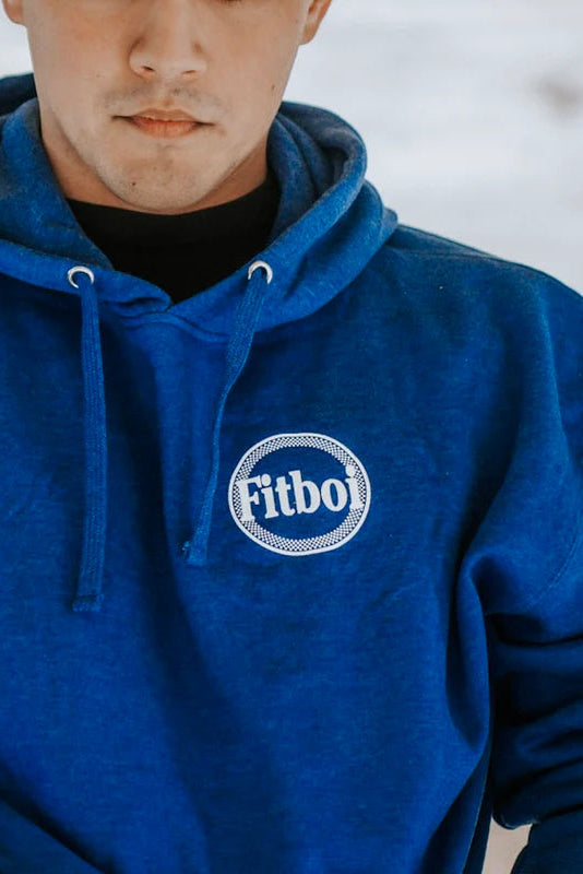 Someone wears the Pillsbury Fitboi - Hoodie by Tango Charlie Apparel, featuring royal blue fabric, white drawstrings, and a round Fitbo logo on the chest, standing out against a snowy, blurred background.