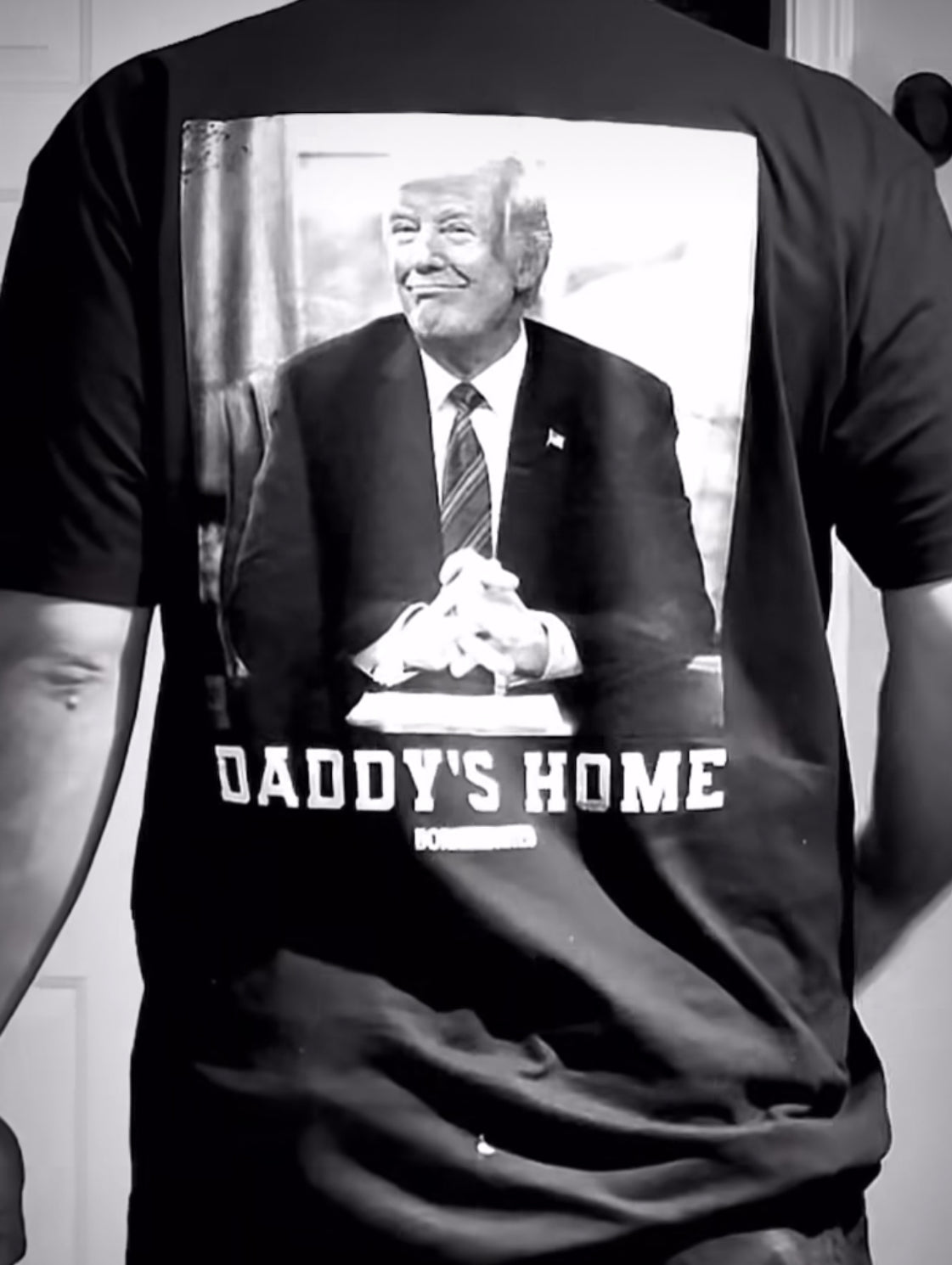 A person is wearing the Daddys Home - #47 - Black premium tee from Born United Private Label, showcasing a striking black and white image of a man sitting with clasped hands and the bold caption DADDYS HOME on the back.