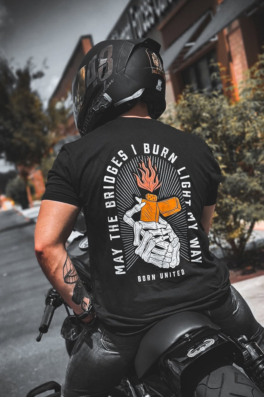 A person in a helmet and a black Burning Bridges (Warehouse) tee by Born United Private Label, featuring skeleton hands shaking over burning dice with the text May the bridges I burn light my way. Born United, rides a motorcycle past a brick building.