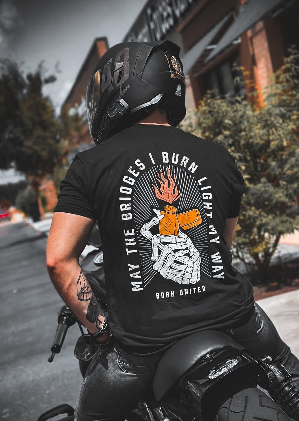 A person in a helmet and a black Burning Bridges (Warehouse) tee by Born United Private Label, featuring skeleton hands shaking over burning dice with the text May the bridges I burn light my way. Born United, rides a motorcycle past a brick building.