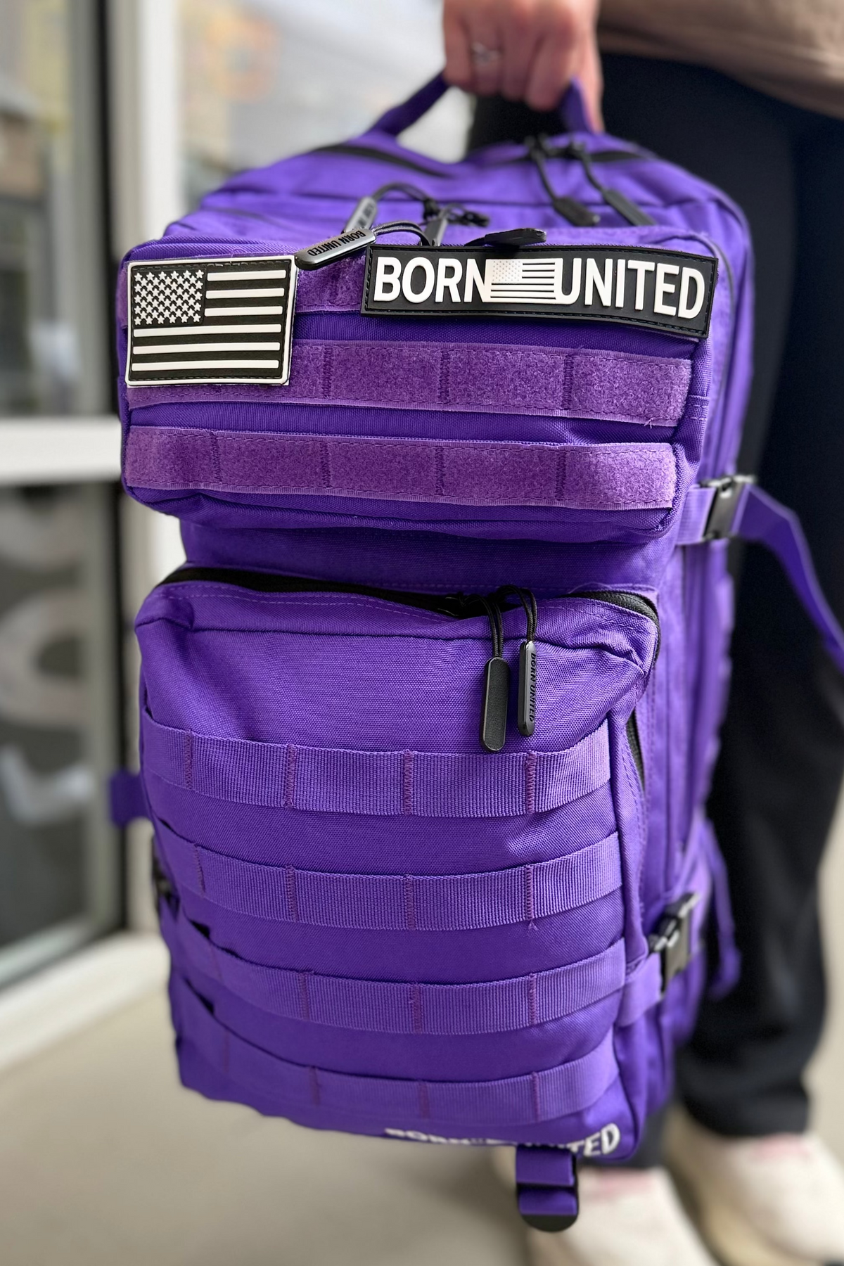 A person holds the 45L Cosmic Purple Tac-Pac backpack from Born United Private Label, featuring an American flag patch and a “BORN UNITED” label. A window and part of the person’s legs are visible in the background.