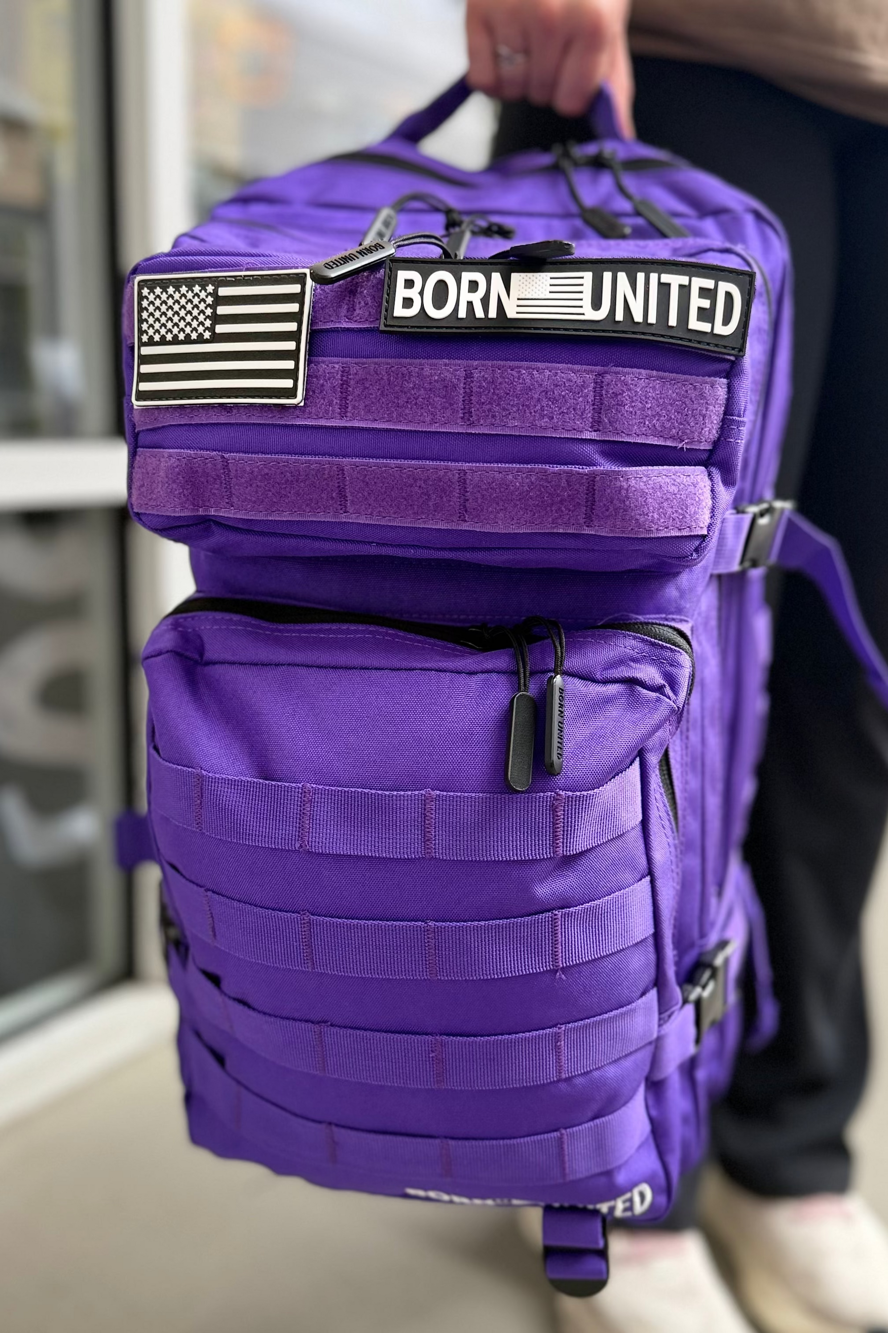 A person holds the 45L Cosmic Purple Tac-Pac backpack from Born United Private Label, featuring an American flag patch and a “BORN UNITED” label. A window and part of the person’s legs are visible in the background.