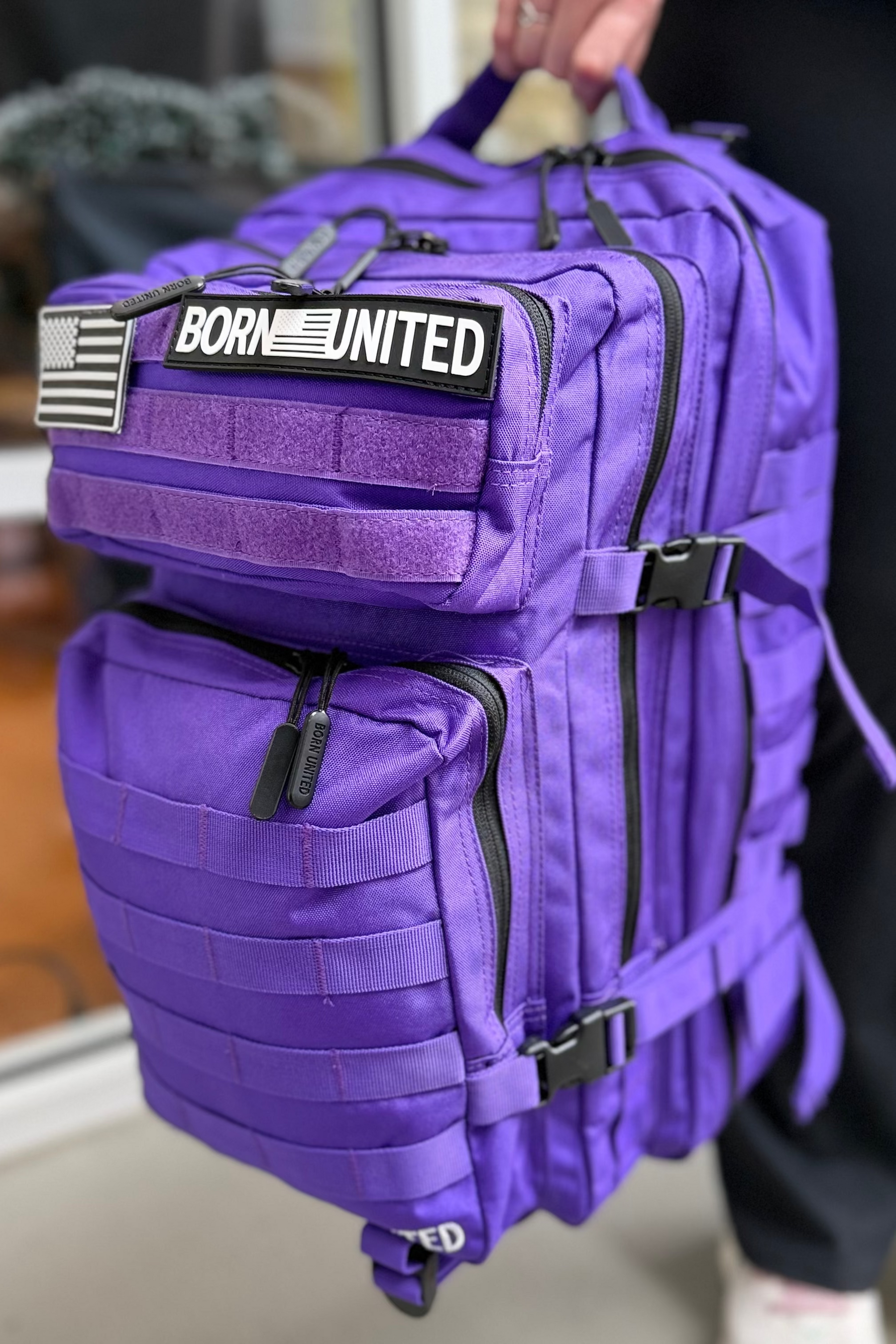 A person holds the Born United Private Label Cosmic Purple Tac-Pac - 45L backpack with black zippers, multiple compartments, and a "BORN UNITED" patch next to a black-and-white American flag patch.