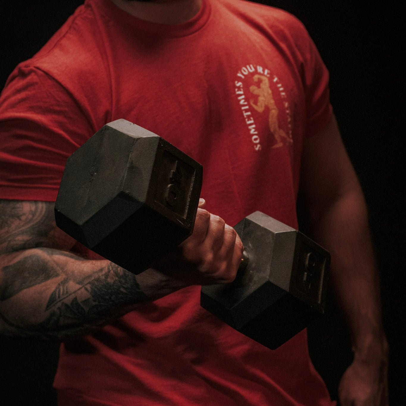 Wearing the "St. Arnold The Stoic" shirt by Tango Charlie Apparel, a tattooed person performs a bicep curl with a large hexagonal dumbbell, upper body visible against a dark background.