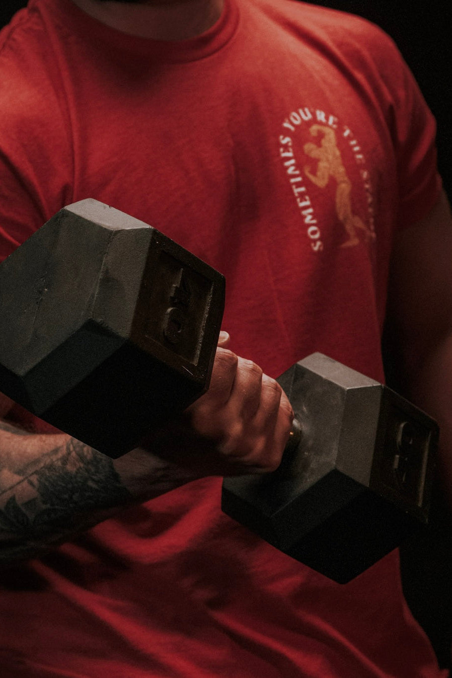 Wearing the "St. Arnold The Stoic" shirt by Tango Charlie Apparel, a tattooed person performs a bicep curl with a large hexagonal dumbbell, upper body visible against a dark background.