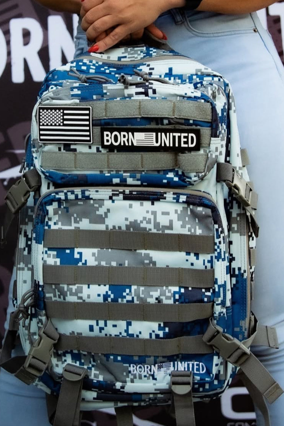 In front of a BORN UNITED backdrop, someone displays the Arctic Tac-Pac - 45L in blue digital camouflage by Born United Private Label. Dressed in light blue jeans, a black top, and wearing a smartwatch, they highlight this products blend of Arctic resilience and adventure-driven unity.