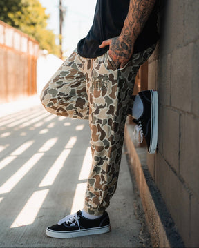 A tattooed person leans against a wall outdoors, wearing Mucker Bucket Men's Classic Premium Joggers in Duck Camo and black Vans. Sunlight filters through a fence, casting shadows on the sidewalk. The person's face is hidden.