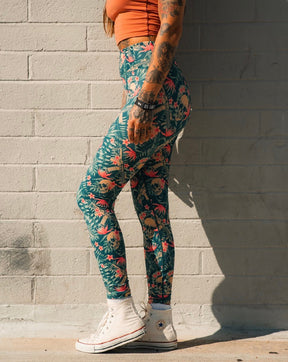 A person with long hair and tattoos, wearing a sleeveless orange crop top and colorful Tango Charlie Apparel Women's Performance Leggings | Aloha AR, plus white socks and high-top sneakers, stands sideways against a light brick wall.
