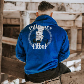 A person sits with their back to the camera, wearing the Tango Charlie Apparel "Pillsbury Fitboi - Hoodie," a bright blue hoodie featuring a cartoon chef lifting weights and the text "Pillsbury Fitboi" in a rustic wooden setting.