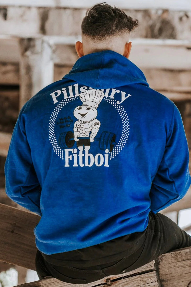 A person sits with their back to the camera, wearing the Tango Charlie Apparel "Pillsbury Fitboi - Hoodie," a bright blue hoodie featuring a cartoon chef lifting weights and the text "Pillsbury Fitboi" in a rustic wooden setting.