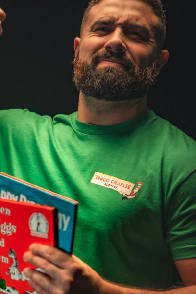 A bearded, tattooed man flexes his bicep to showcase strength while holding Dr. Seuss books: Green Eggs and Ham and Happy Birthday to You! He wears a green St. Seuss Tee from Tango Charlie Apparel with a red name tag on the chest.