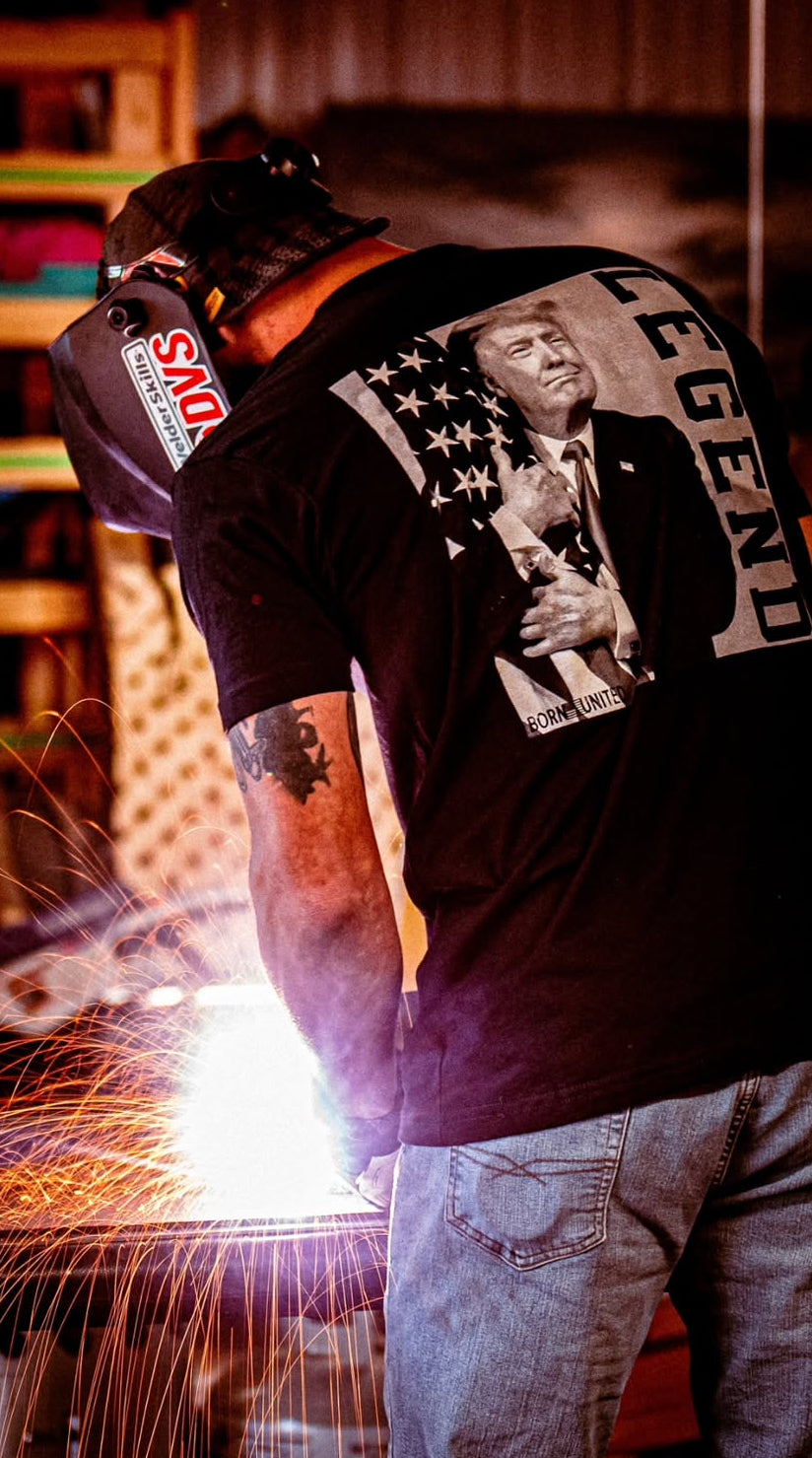 Welding in a protective helmet, a person sports a Born United Private Label black T-shirt labeled Legend - Trump, depicting the president holding a cat against an American flag. Bright sparks fly.