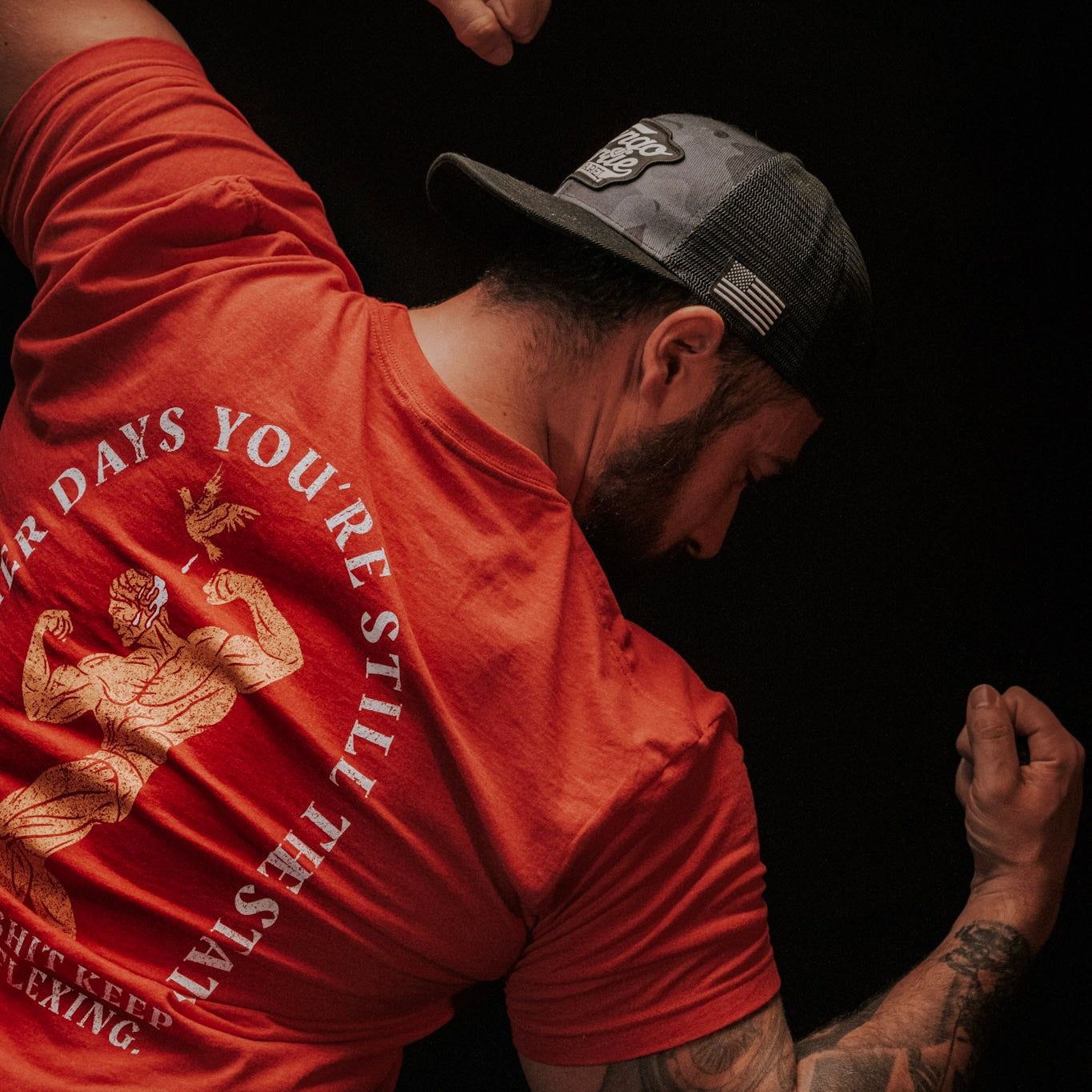 A man flexes his arm muscles in a Tango Charlie Apparel "St. Arnold The Stoic" t-shirt and a backward black cap. The shirt’s back shows an illustration with the phrase: "After days you’re still the last one flexing.
