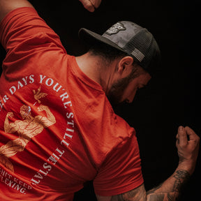A man flexes his arm muscles in a Tango Charlie Apparel "St. Arnold The Stoic" t-shirt and a backward black cap. The shirt’s back shows an illustration with the phrase: "After days you’re still the last one flexing.