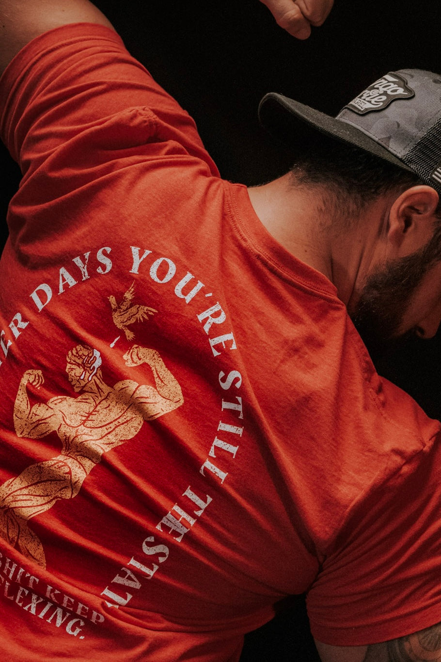 A man flexes his arm muscles in a Tango Charlie Apparel "St. Arnold The Stoic" t-shirt and a backward black cap. The shirt’s back shows an illustration with the phrase: "After days you’re still the last one flexing.