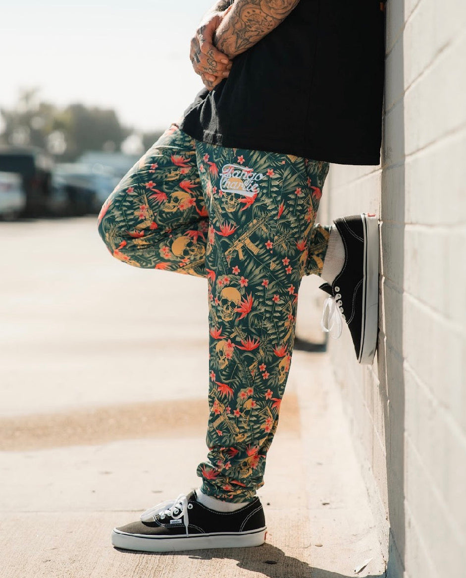 A tattooed person stands outdoors against a light brick wall, wearing Tango Charlie Apparel’s Men's Classic Premium Joggers | Aloha AR with a tropical pattern—perfect for running—paired with a black shirt and black sneakers. Cropped from the shoulders up.