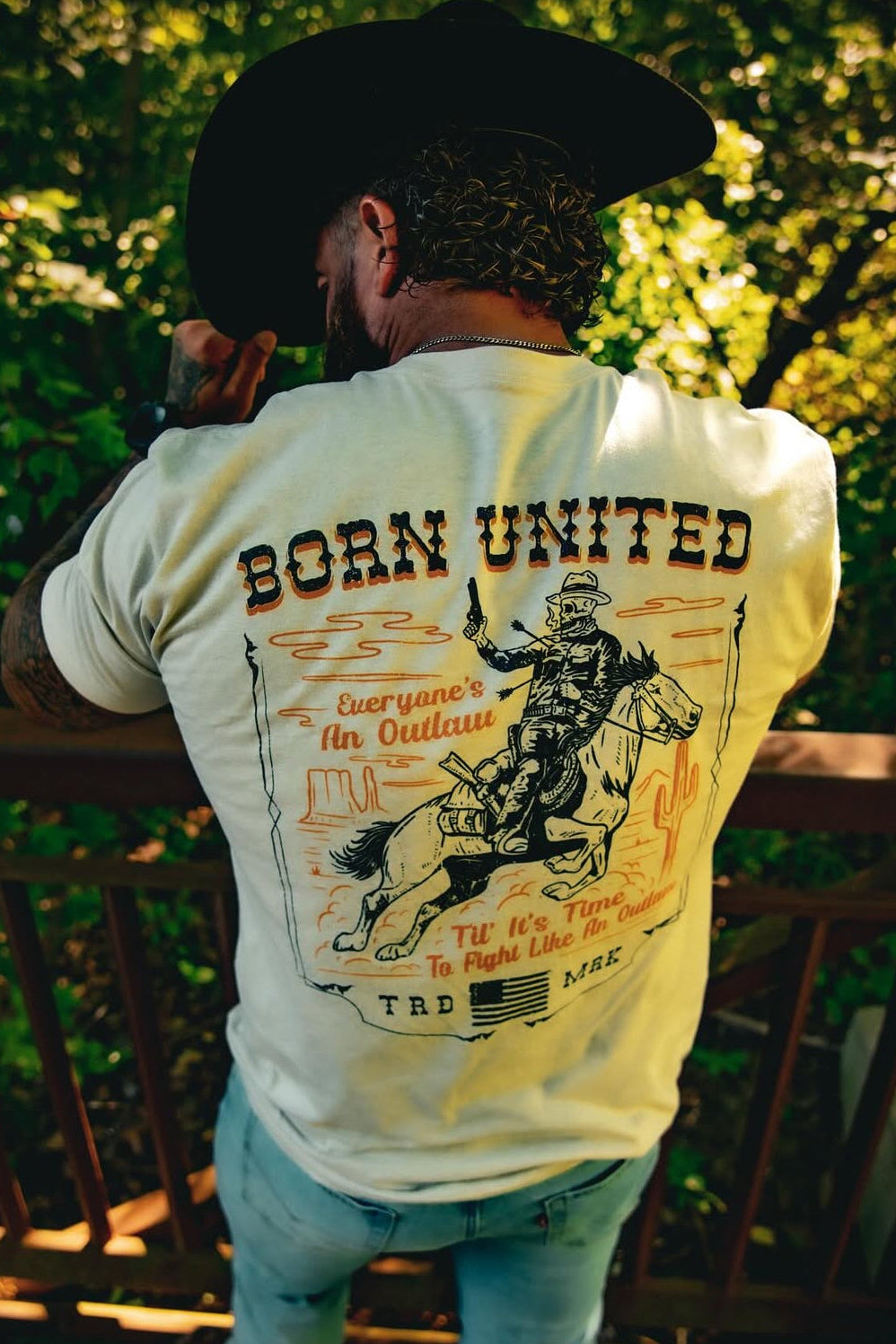 A man in a cowboy hat stands on a wooden deck, wearing the Born United Private Labels Outlaw Tee in tan. The tee features a cowboy on horseback with the phrase Everyones an Outlaw. Lush green foliage provides a fitting backdrop for this rugged scene.