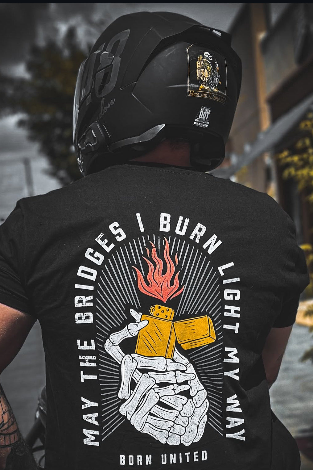 A person wearing a helmet and a black Burning Bridges (Warehouse) tee from Born United Private Label, showcasing a skeleton hand with a flaming lighter. The text reads, MAY THE BRIDGES I BURN LIGHT MY WAY. BORN UNITED, with a building in lush greenery as the backdrop.