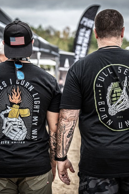 At an outdoor event, two men sport bold black T-shirts. The left shirt reads May the Bridges I Burn Light My Way with subtle flames from Burning Bridges (Warehouse), while the right features Full Send Born United, emphasizing its originality as a Born United Private Label iconic tee.