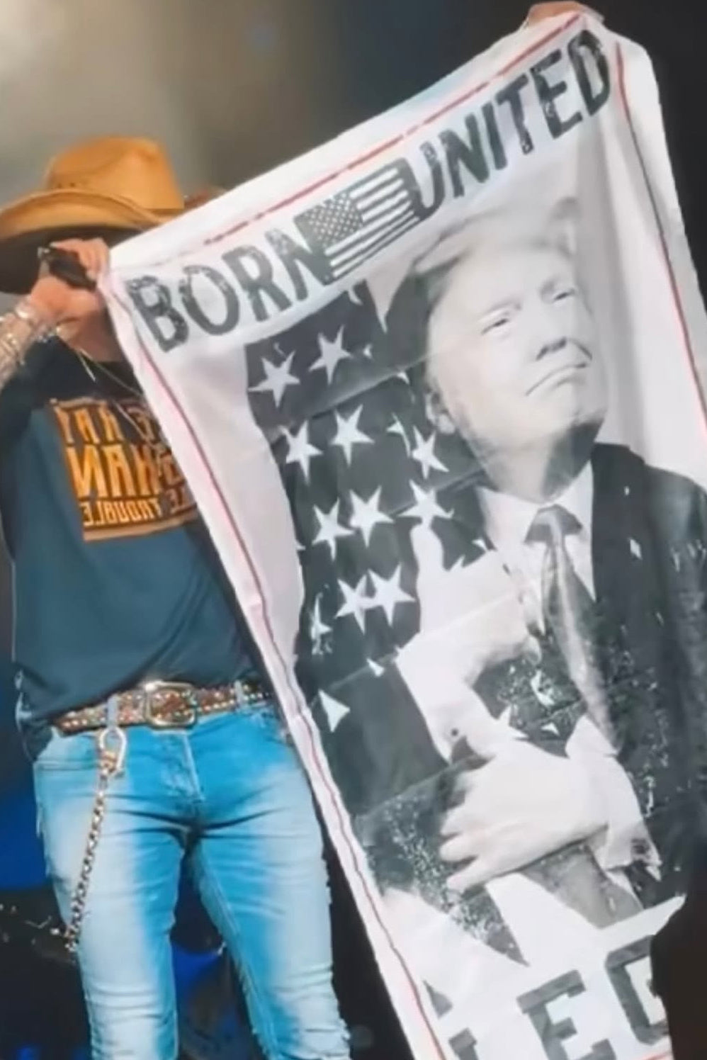 A person in a wide-brimmed hat, jeans, and a T-shirt displays the Legend Trump Flag from Born United Private Label, showcasing President Donald Trump against an American flag with the words Born United, embodying the 2024 election spirit.