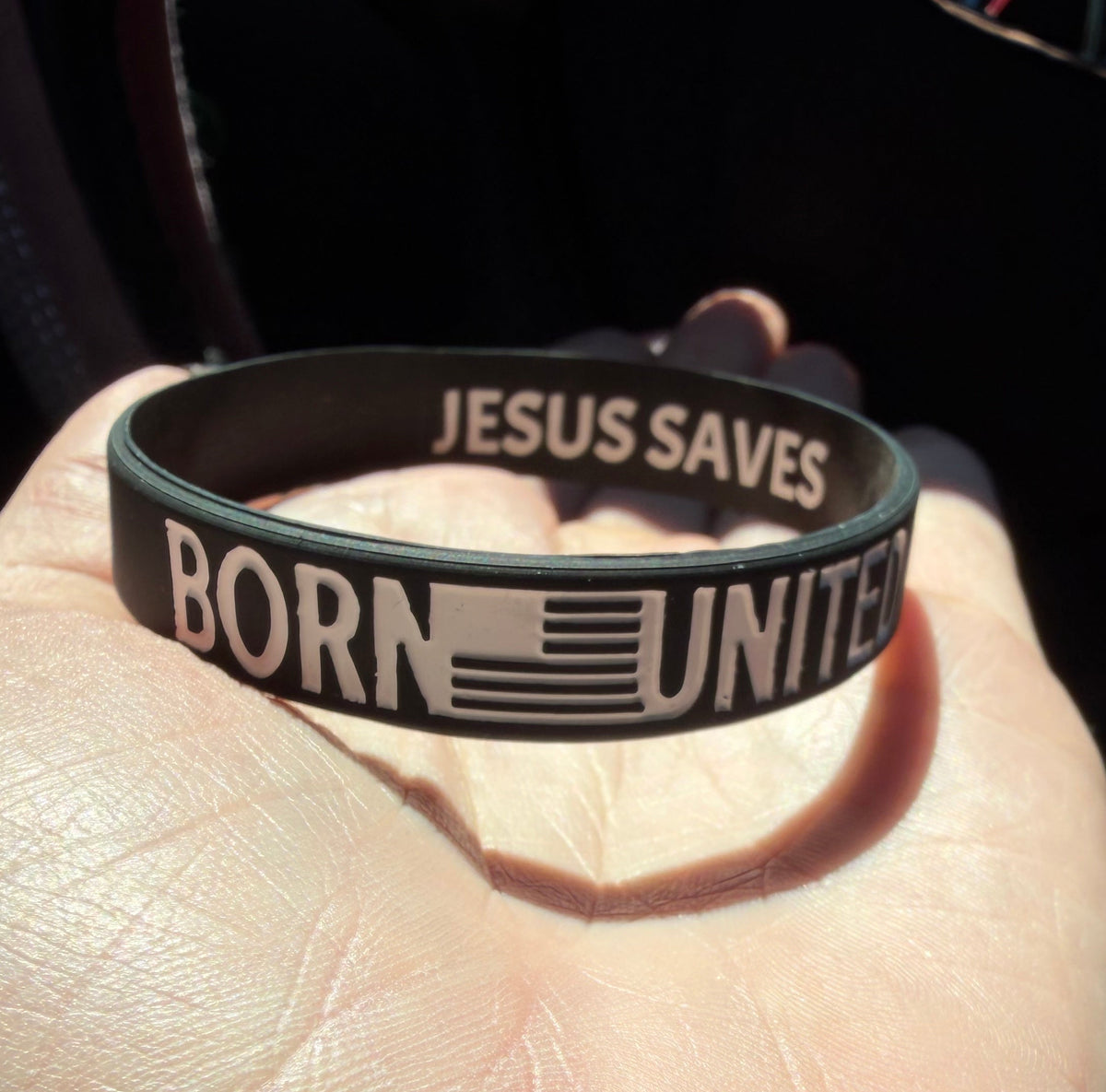 A hand holds the Born United Bracelet by Born United Private Label, a black silicone wristband with "BORN UNITED" and an American flag on the front, and "JESUS SAVES" printed inside.