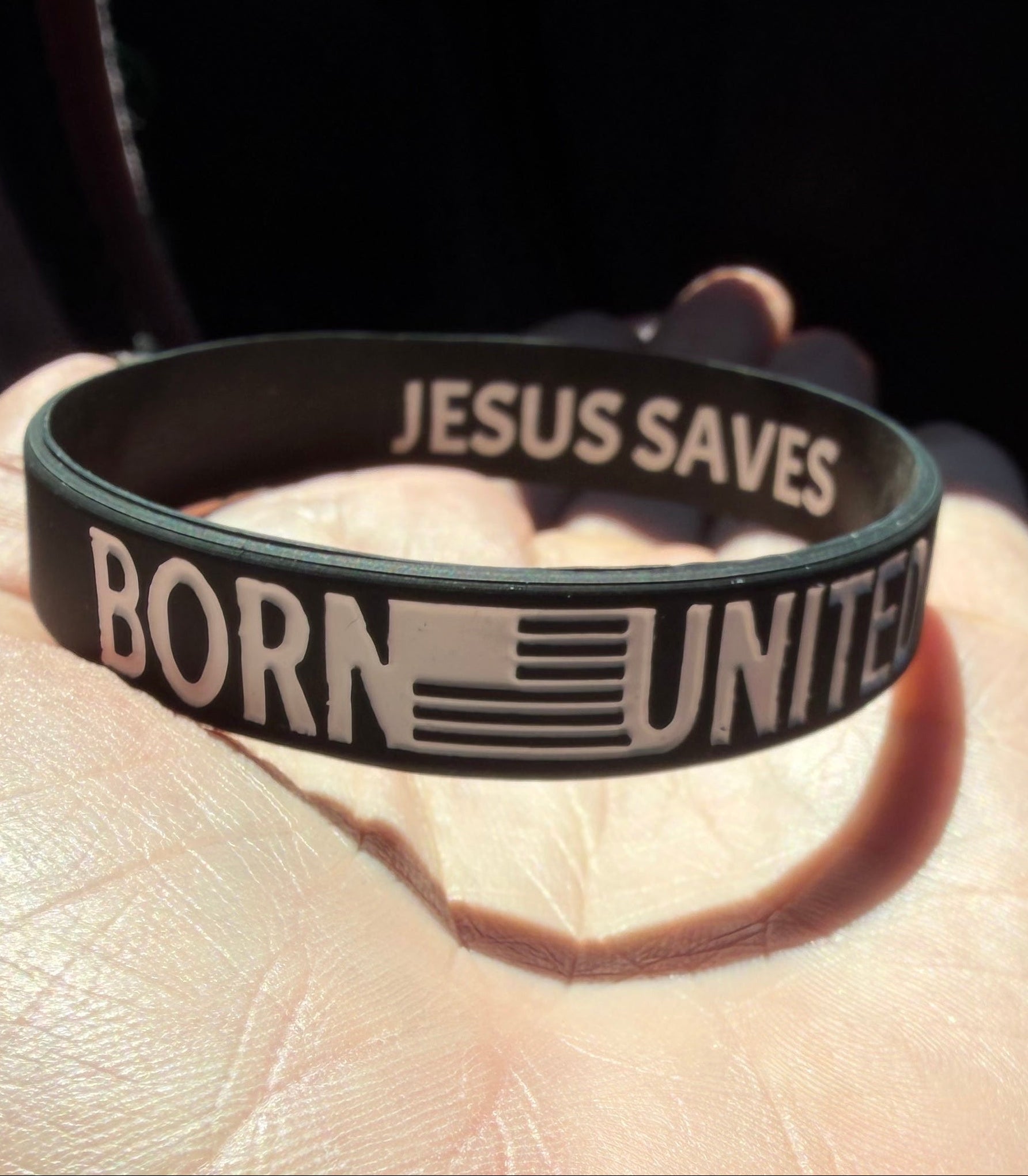 A hand holds the Born United Bracelet by Born United Private Label, a black silicone wristband with "BORN UNITED" and an American flag on the front, and "JESUS SAVES" printed inside.