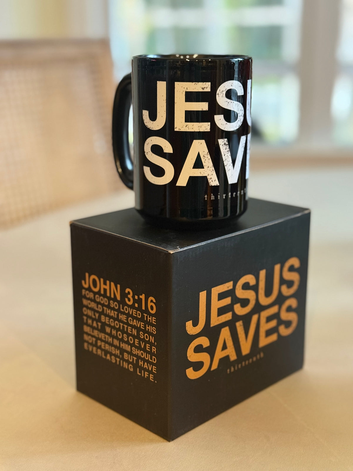 The 13th Jesus Saves Mug features bold white text and comes in a matching black box with JESUS SAVES and John 3:16 in gold. A softly blurred background highlights its faith-inspired design.