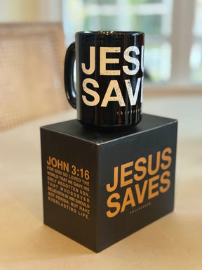 The 13th Jesus Saves Mug features bold white text and comes in a matching black box with JESUS SAVES and John 3:16 in gold. A softly blurred background highlights its faith-inspired design.