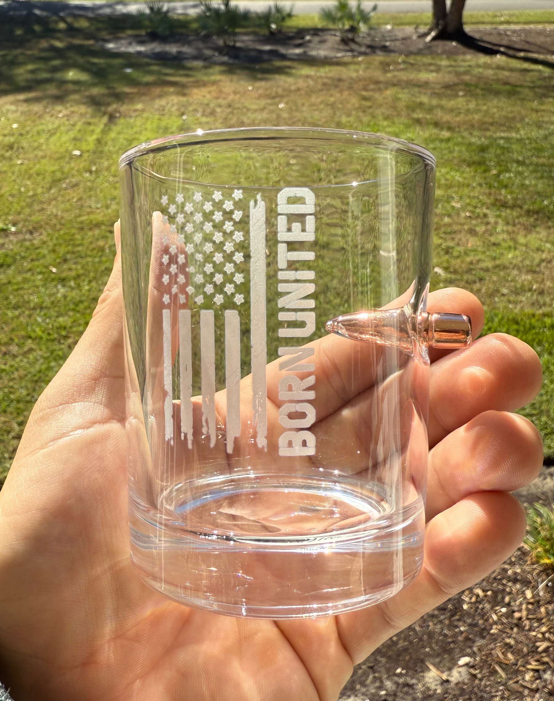 A hand holds the Born United Bullet Whiskey Glass by Born United Private Label, featuring an engraved American flag, "BORN UNITED," and a real bullet embedded in the side. Sunlit grass, trees, and houses appear in the background.