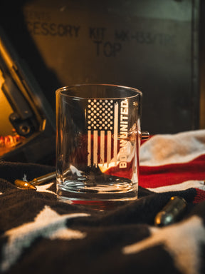 The Born United Bullet Whiskey Glass by Born United Private Label features an embedded bullet and American flag design, displayed on a folded flag with scattered bullets and military-style text in the dim background.