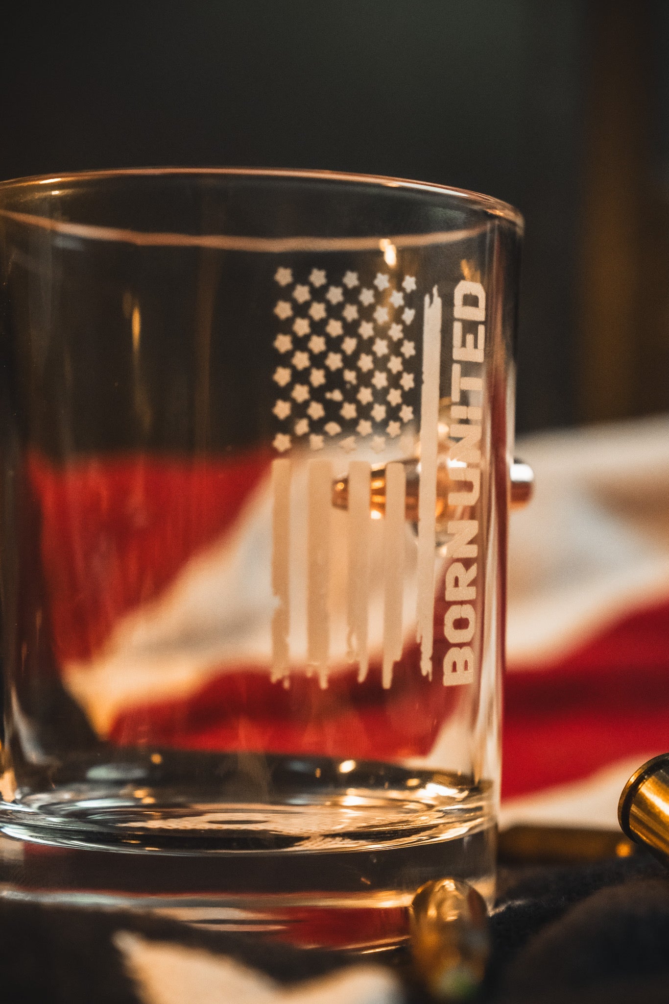 The Born United Bullet Whiskey Glass by Born United Private Label features an etched American flag and "Born United" text, shown on a surface with a blurred American flag and bullet casings in the background.