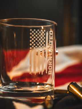 The Born United Bullet Whiskey Glass by Born United Private Label features an etched American flag and "Born United" text, shown on a surface with a blurred American flag and bullet casings in the background.