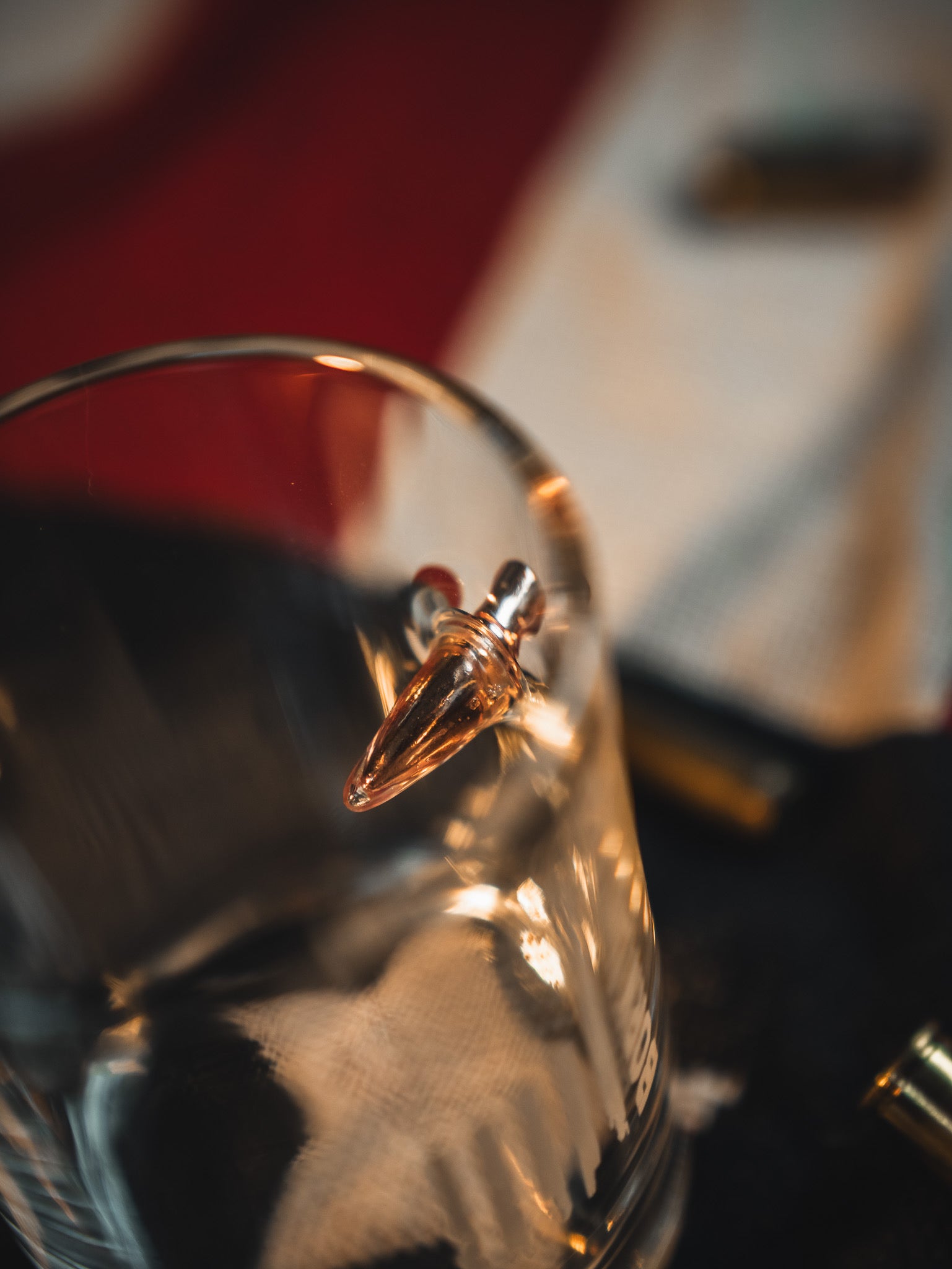 A close-up of the Born United Bullet Whiskey Glass by Born United Private Label, showcasing a bullet embedded in its side against a blurred red, white, and black backdrop.