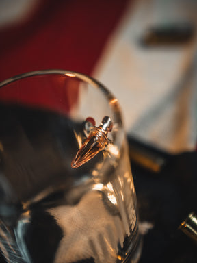 A close-up of the Born United Bullet Whiskey Glass by Born United Private Label, showcasing a bullet embedded in its side against a blurred red, white, and black backdrop.
