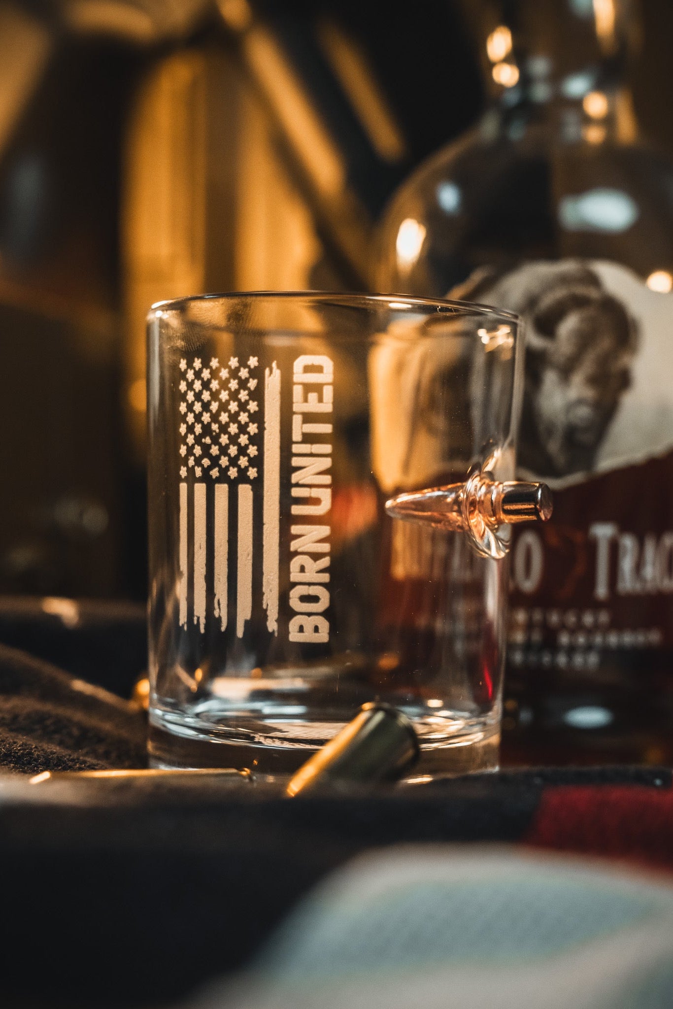The Born United Bullet Whiskey Glass by Born United Private Label, featuring a stylized American flag, "BORN UNITED" text, and an embedded bullet, sits beside a Buffalo Trace bourbon bottle with bullet casings and a flag-patterned cloth in front.
