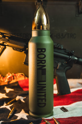 A Bullet Thermos from Born United Private Label stands upright on a U.S. flag, surrounded by rifle bullets with a rifle in the background under warm lighting.