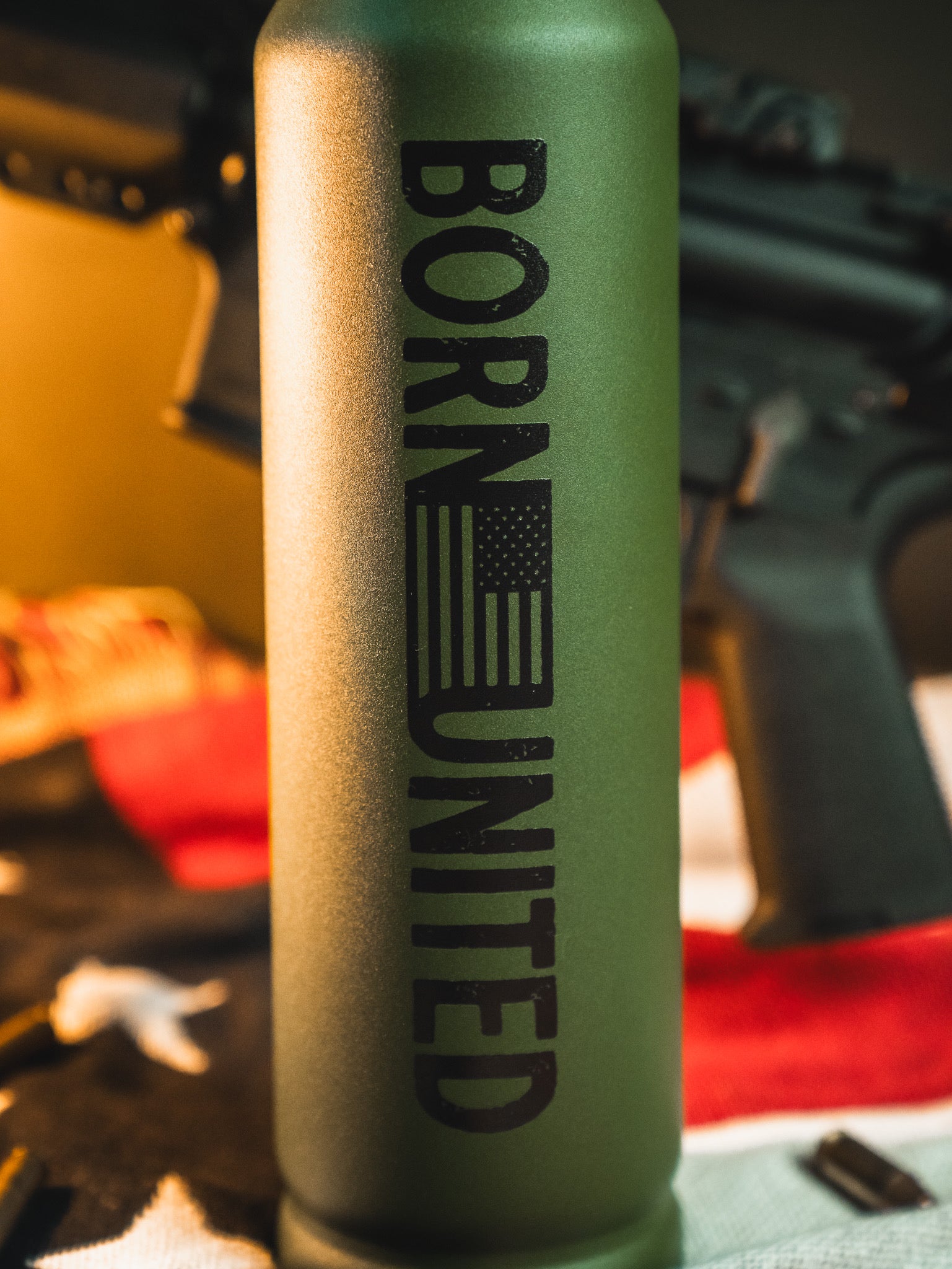 A green Bullet Thermos by Born United Private Label features the text BORN UNITED and a vertical American flag. The background includes rifle parts, a blurred U.S. flag, and scattered bullet casings.