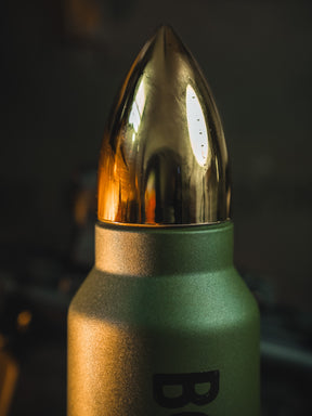 A close-up of a green metallic Bullet Thermos by Born United Private Label with a shiny, gold bullet-shaped cap reflecting light against a dark, blurred background.
