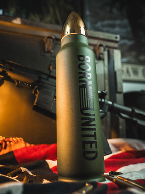 A green Bullet Thermos from Born United Private Label, featuring an American flag design and BORN UNITED text, sits on a surface with a blurred rifle and flag in the background.