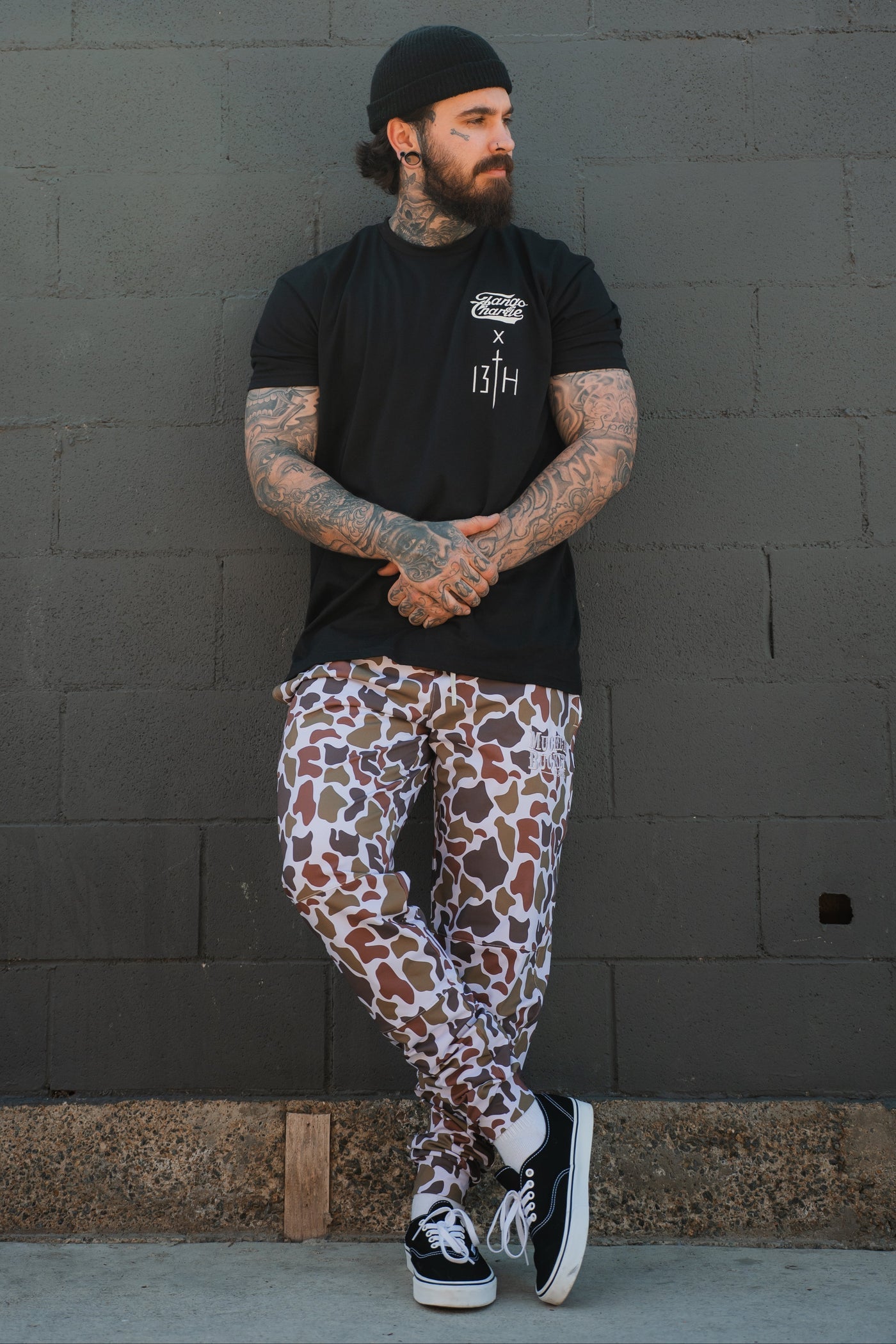 A tattooed, bearded man in a black beanie leans against a gray brick wall wearing a black T-shirt, Mucker Bucket Men's Classic Premium Joggers in Old School Duck Camo, and black Vans sneakers with white soles.