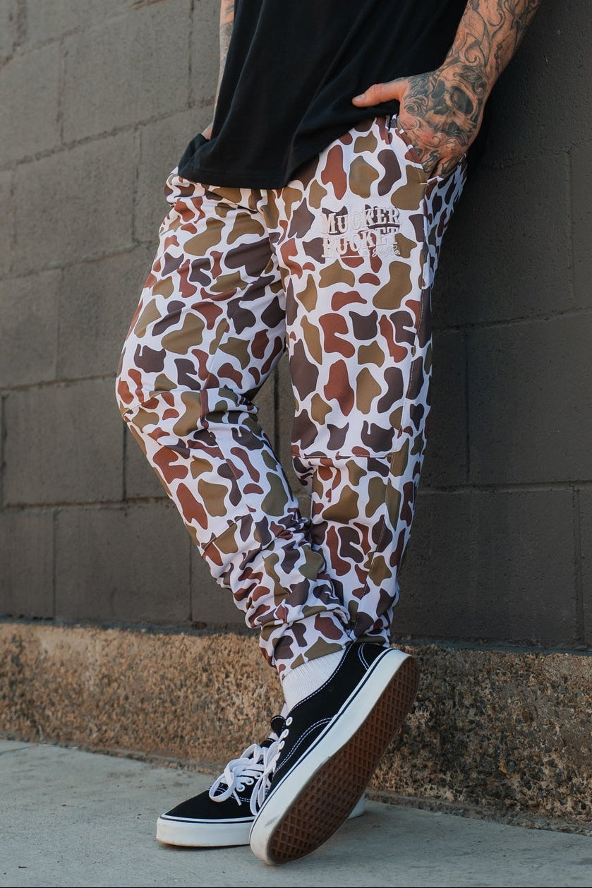 A bearded man in a black beanie, black tee, and Mucker Bucket Men's Classic Premium Joggers in Old School Duck Camo stands by a gray brick wall, showing tattooed arms with one hand in his pocket. He finishes the look with black-and-white sneakers.