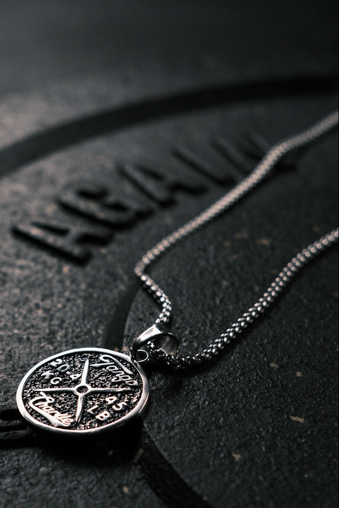 A Barbell Necklace by Tango Charlie Apparel lies on a textured dark surface with AGAIN partially visible in embossed letters. The focus is on its elegant and intricate design, combining reliability with a lifetime warranty.