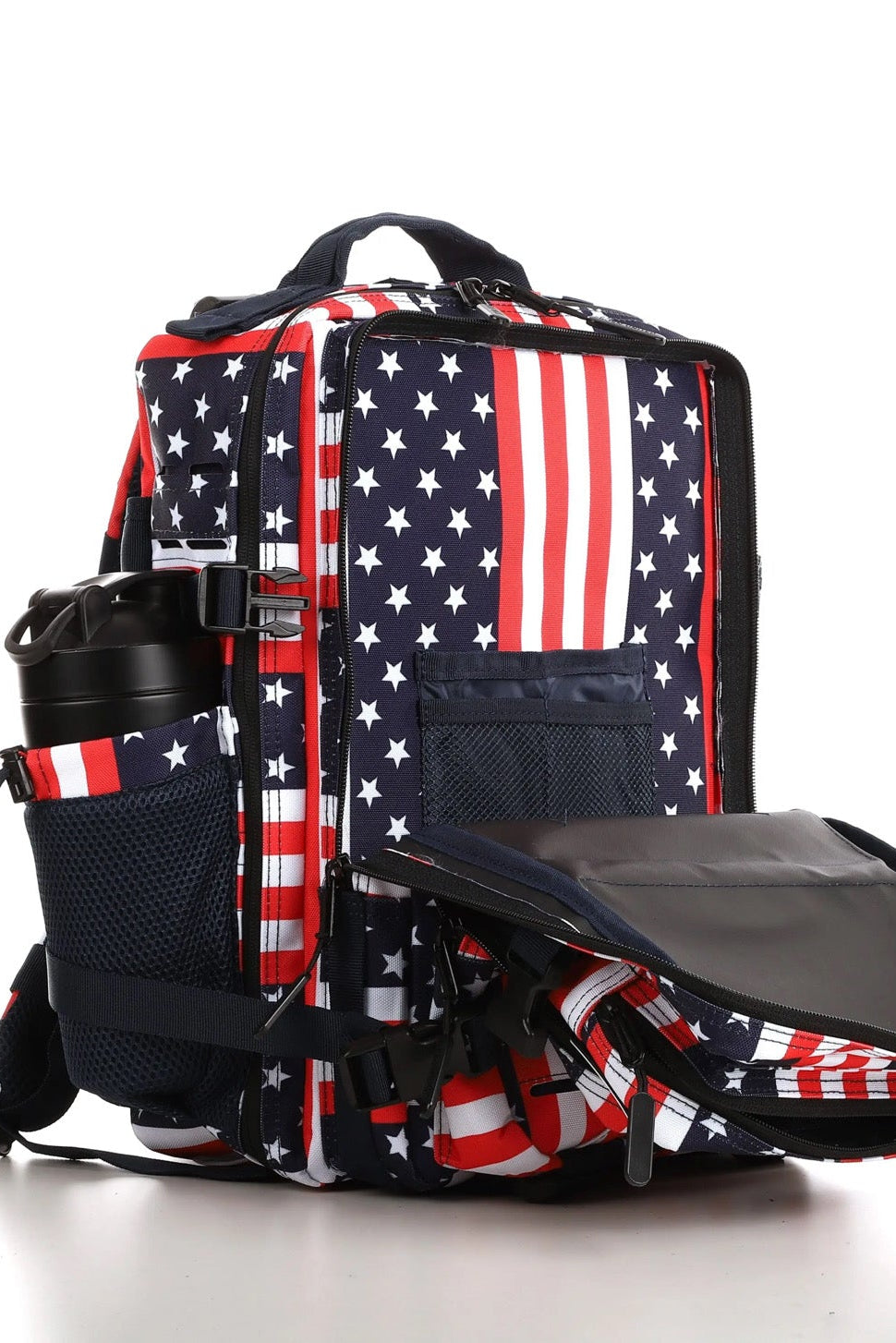 The Ultimate Patriot Special Edition Tac-Pac - 25L by Born United Private Label features an American flag design with red and white stripes and stars on blue. It has multiple compartments, including a side pocket with a black thermos, all showcased on a reflective white surface.