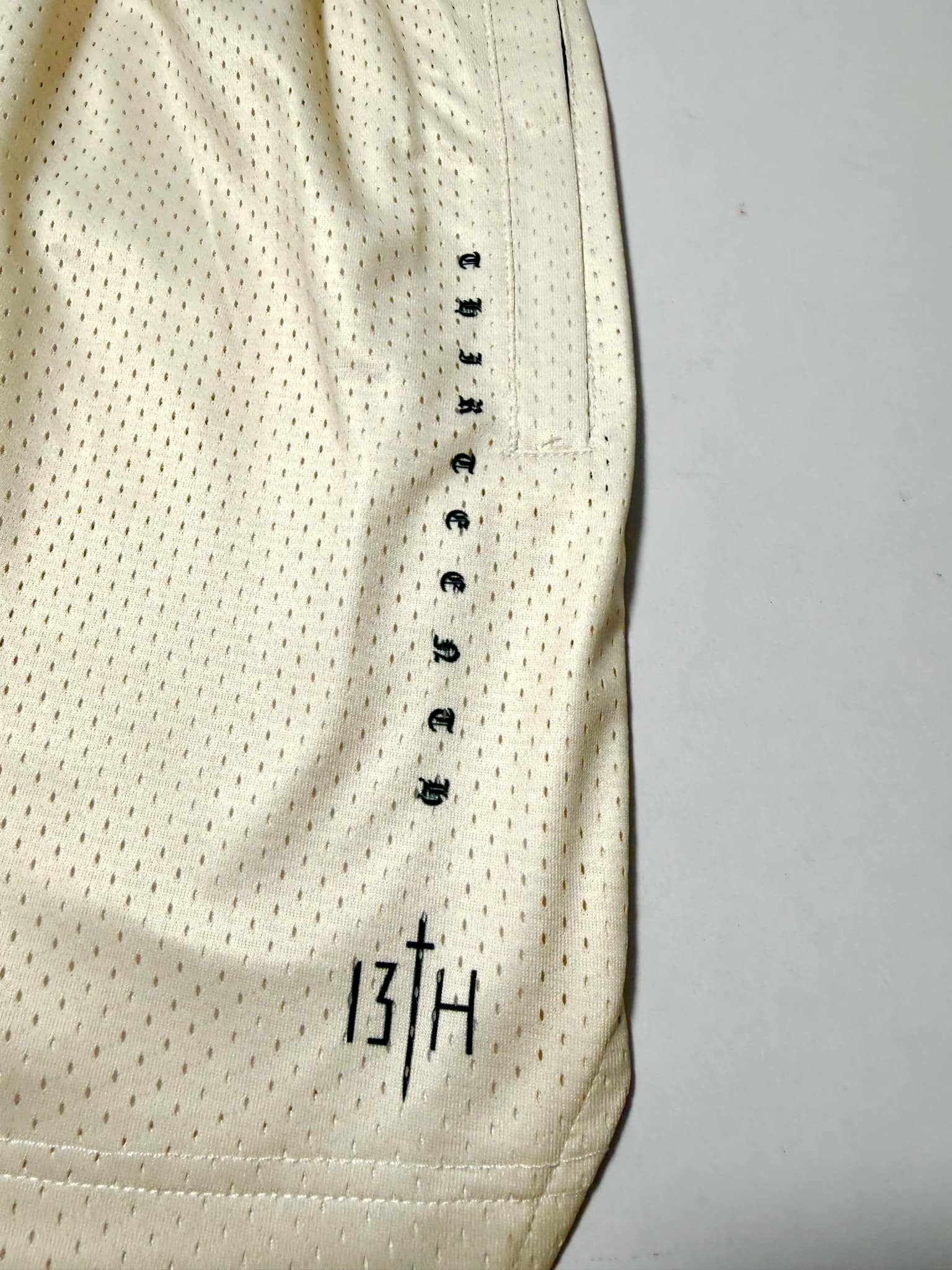 Introducing the Men’s Mesh Lifestyle Shorts | 5.5" – Christ is King Ivory by 13th. These unisex, cream-colored mesh shorts feature bold vertical "thirteenth" text and a distinctive black 13TH logo near the hem. Combining comfort and style, they’re perfect for any activity.