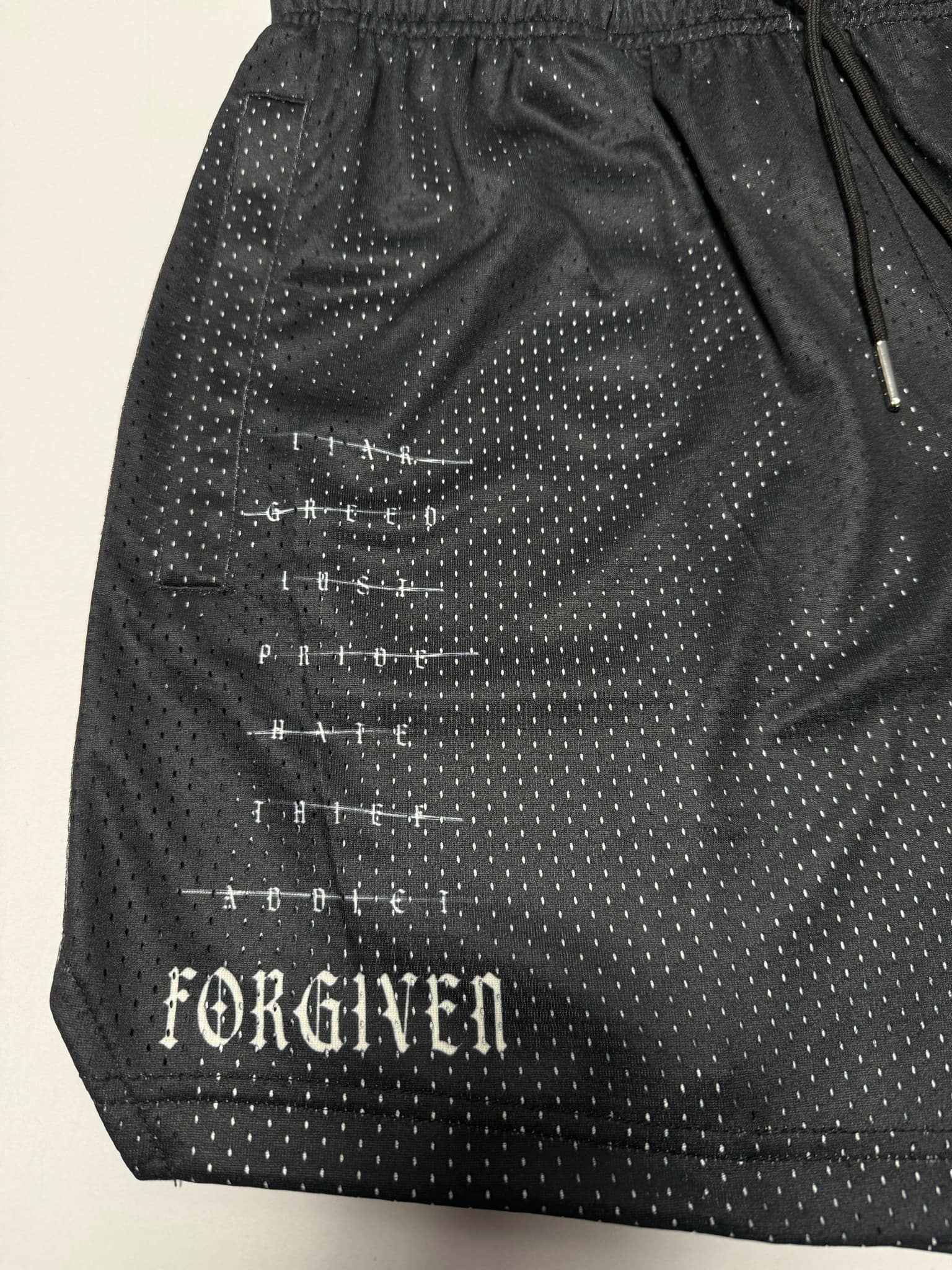 The Men's Mesh Lifestyle Shorts | 5.5" - Forgiven by 13th feature black perforated fabric with crossed-out words such as LIAR, GREED, LUST, PRIDE, HATE, THIEF, and ADDICT, while FORGIVEN stands out in bold white text at the bottom. These unisex shorts from 13th blend athletic comfort with a powerful statement.