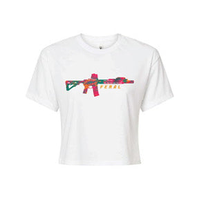 The Feral Crop Tee - White by Tango Charlie Apparel features a colorful floral rifle graphic with "FERAL" in yellow and pink beside it. Made from a soft cotton-poly blend for maximum comfort.