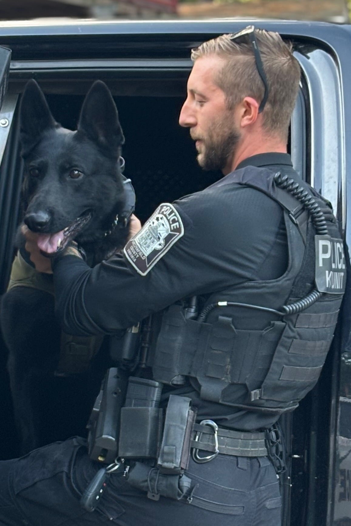 A Chesterfield County Police officer in tactical gear, wearing sunglasses on his head, helps K9 Knight—a black police dog—out of a vehicle while holding the dog’s collar.
