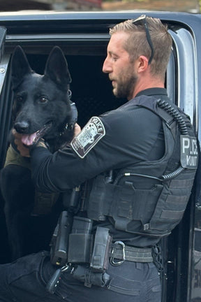 A Chesterfield County Police officer in tactical gear, wearing sunglasses on his head, helps K9 Knight—a black police dog—out of a vehicle while holding the dog’s collar.